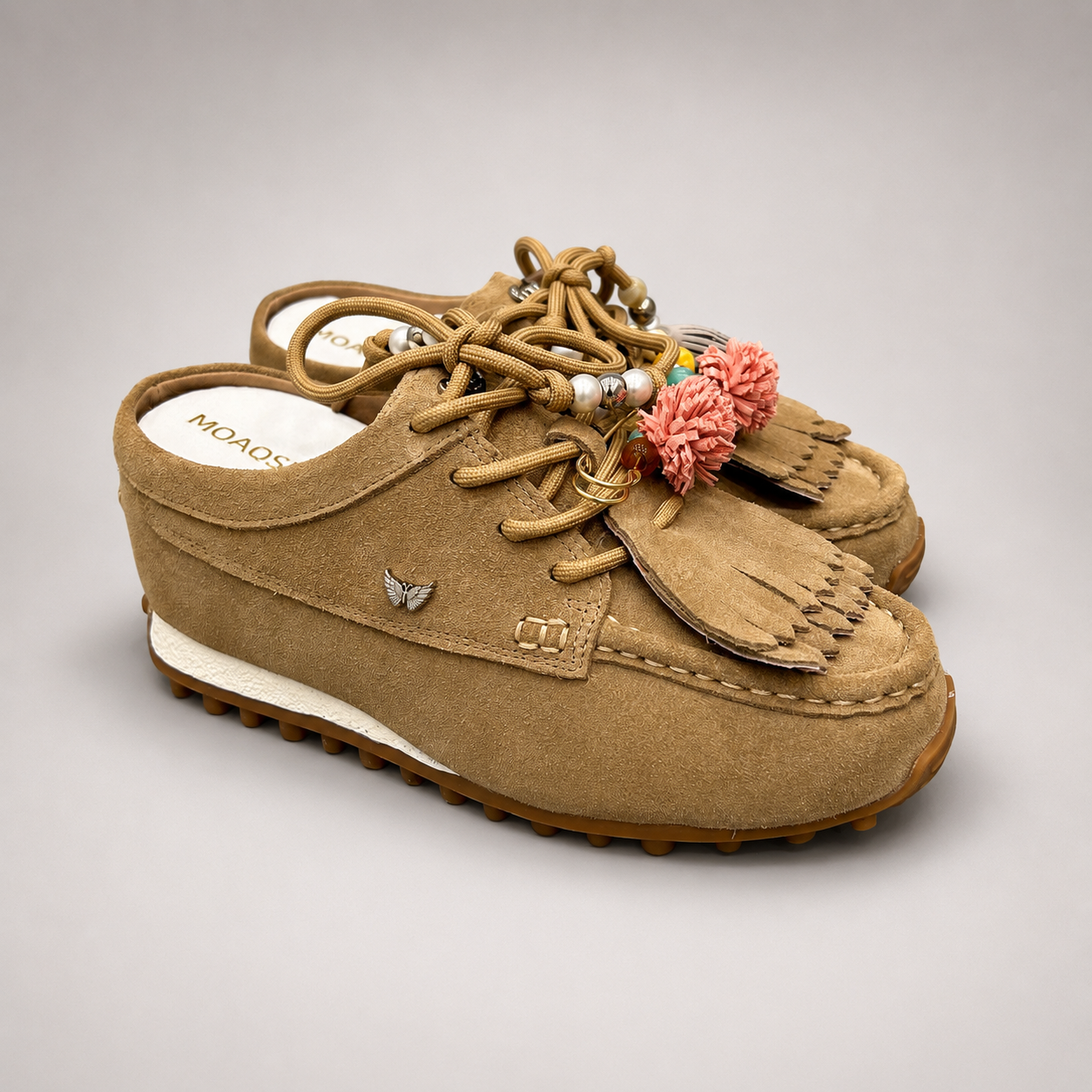 Tan moccasin sneaker pair three-quarter view showing fringe and tassel details over the laces